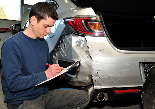 A man in a blue long-sleeved shirt and gray work pants kneels near a damaged gray sedan, holding a clipboard and pen. He is recording information for a collision repair estimate.