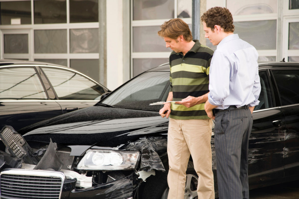 Two men wearing business-casual attire looking at a black sedan with front-end damage. The man in the foreground, a white man with curly brown hair, has his hands in his pockets and is looking at the vehicle, while the man slightly behind him, a white man of similar age with chin-length straight brown hair, is holding paperwork and also looking at the vehicle.